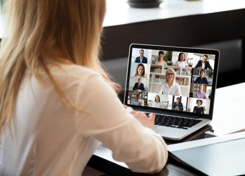 View over businesswoman shoulder sit at workplace desk looks at pc screen during group videocall with different age and ethnicity colleagues using application and webcam, distant communication concept