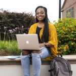 A student using a laptop while sitting on a concrete bench outdoors in front of bushes.