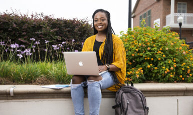 A student using a laptop while sitting on a concrete bench outdoors in front of bushes.