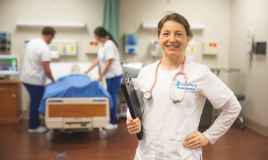 Nursing student with a stethoscope around her neck holds a clipboard in her right hand while two more nursing students work in the background