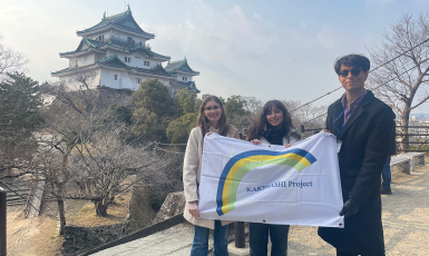 Three students hold a flag that says Kakehashi Project in front of a Japanese temple.