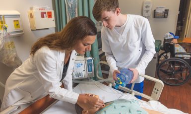 A faculty and student using a bag valve mask on a dummy model laying in a hospital bed in a hospital simulation lab setting.