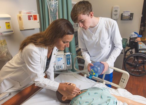 A faculty and student using a bag valve mask on a dummy model laying in a hospital bed in a hospital simulation lab setting.