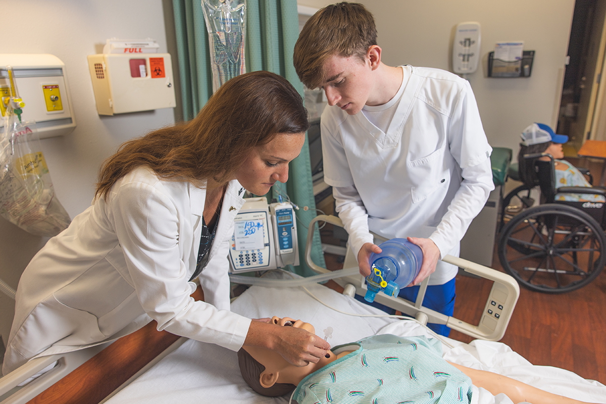 A faculty and student using a bag valve mask on a dummy model laying in a hospital bed in a hospital simulation lab setting.
