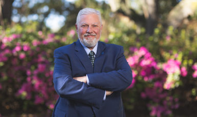 David Brinkley, new executive director of WUWF, poses with arms crossed outside in front of bushes blooming with pink flowers