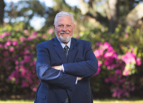 David Brinkley, new executive director of WUWF, poses with arms crossed outside in front of bushes blooming with pink flowers