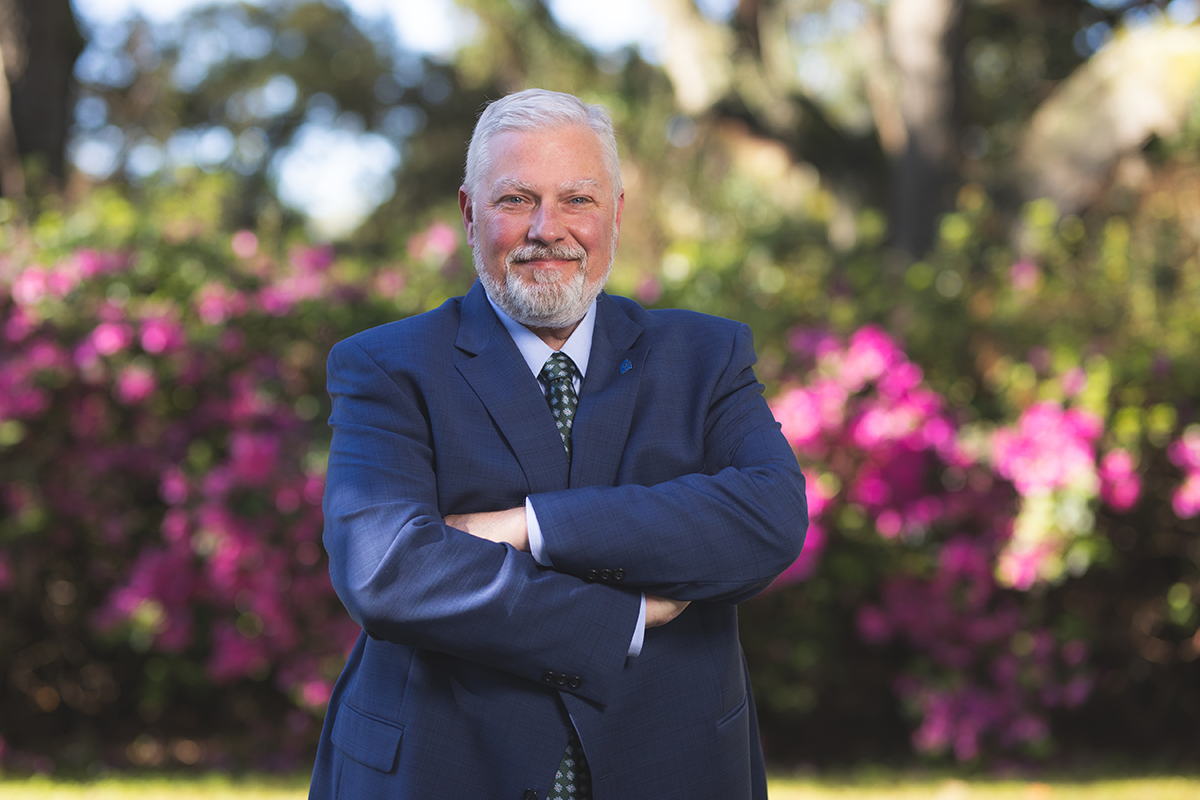 David Brinkley, new executive director of WUWF, poses with arms crossed outside in front of bushes blooming with pink flowers