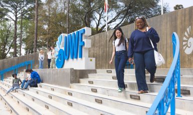 Two students walk down the steps to the right of the UWF entrance sign that is colored blue, while three other students stand to the left of the sign chatting amongst themselves and three more sit on the concrete steps in front of the sign.