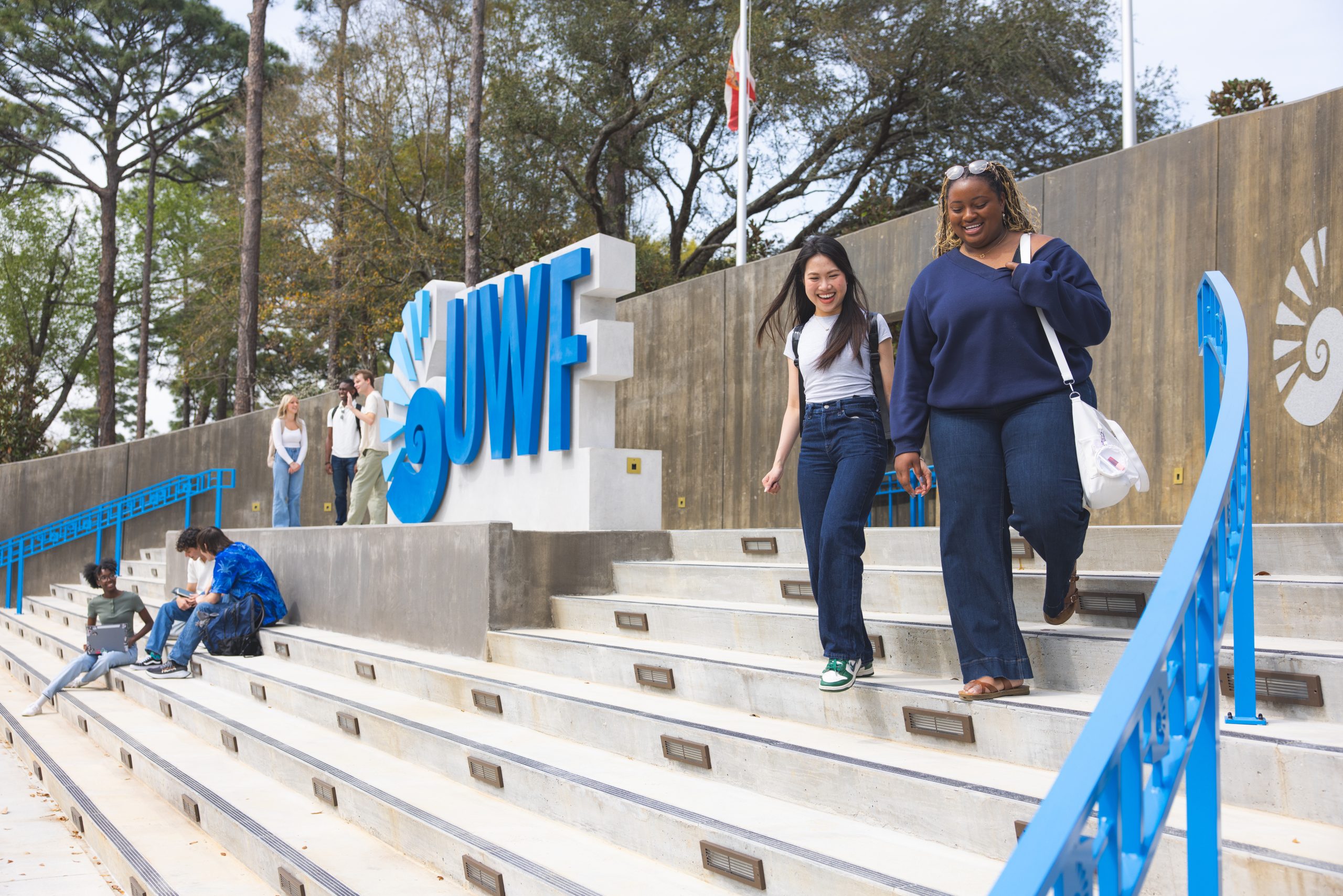 Two students walk down the steps to the right of the UWF entrance sign that is colored blue, while three other students stand to the left of the sign chatting amongst themselves and three more sit on the concrete steps in front of the sign.
