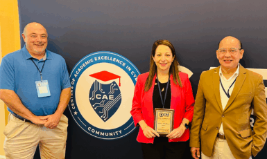 Three people stand in front of a wall around a logo that reads, "Centers of Academic Excellence in Cybersecurity Community. One holds a plaque award in her hands.