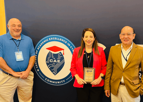 Three people stand in front of a wall around a logo that reads, "Centers of Academic Excellence in Cybersecurity Community. One holds a plaque award in her hands.