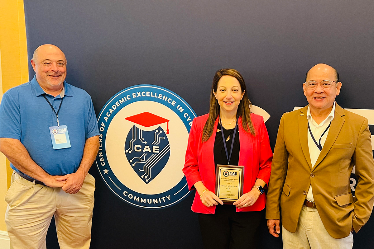 Three people stand in front of a wall around a logo that reads, "Centers of Academic Excellence in Cybersecurity Community. One holds a plaque award in her hands.