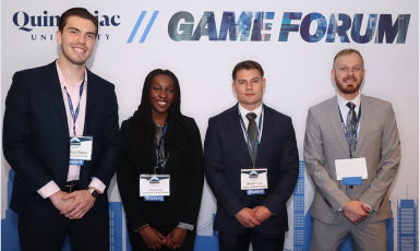 Four students stand smiling in front of a colored-white backdrop that says, "Quinnipiac University Game Forum"