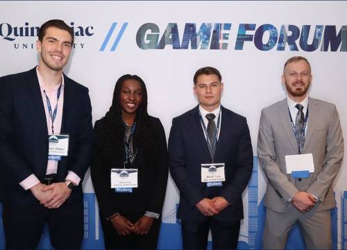 Four students stand smiling in front of a colored-white backdrop that says, "Quinnipiac University Game Forum"