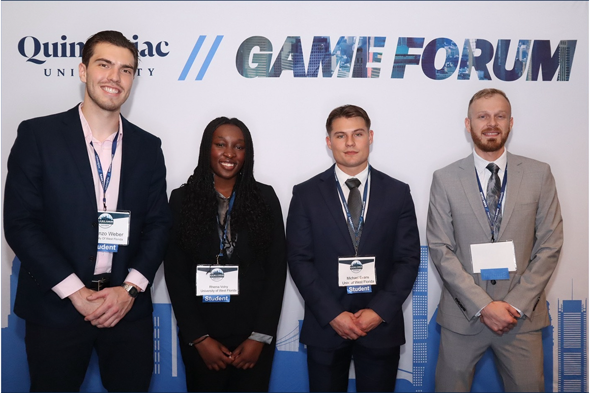 Four students stand smiling in front of a colored-white backdrop that says, "Quinnipiac University Game Forum"