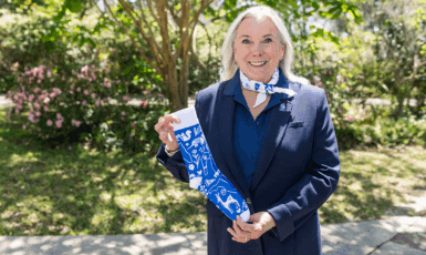 UWF President Martha Saunders stands smiling with a colored-white bandana tied around her neck and holding a colored-blue and white sock in her hands.