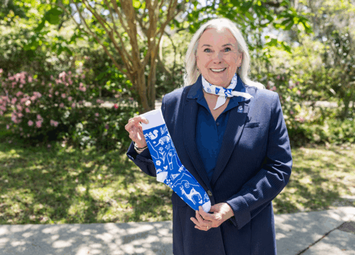 UWF President Martha Saunders stands smiling with a colored-white bandana tied around her neck and holding a colored-blue and white sock in her hands.