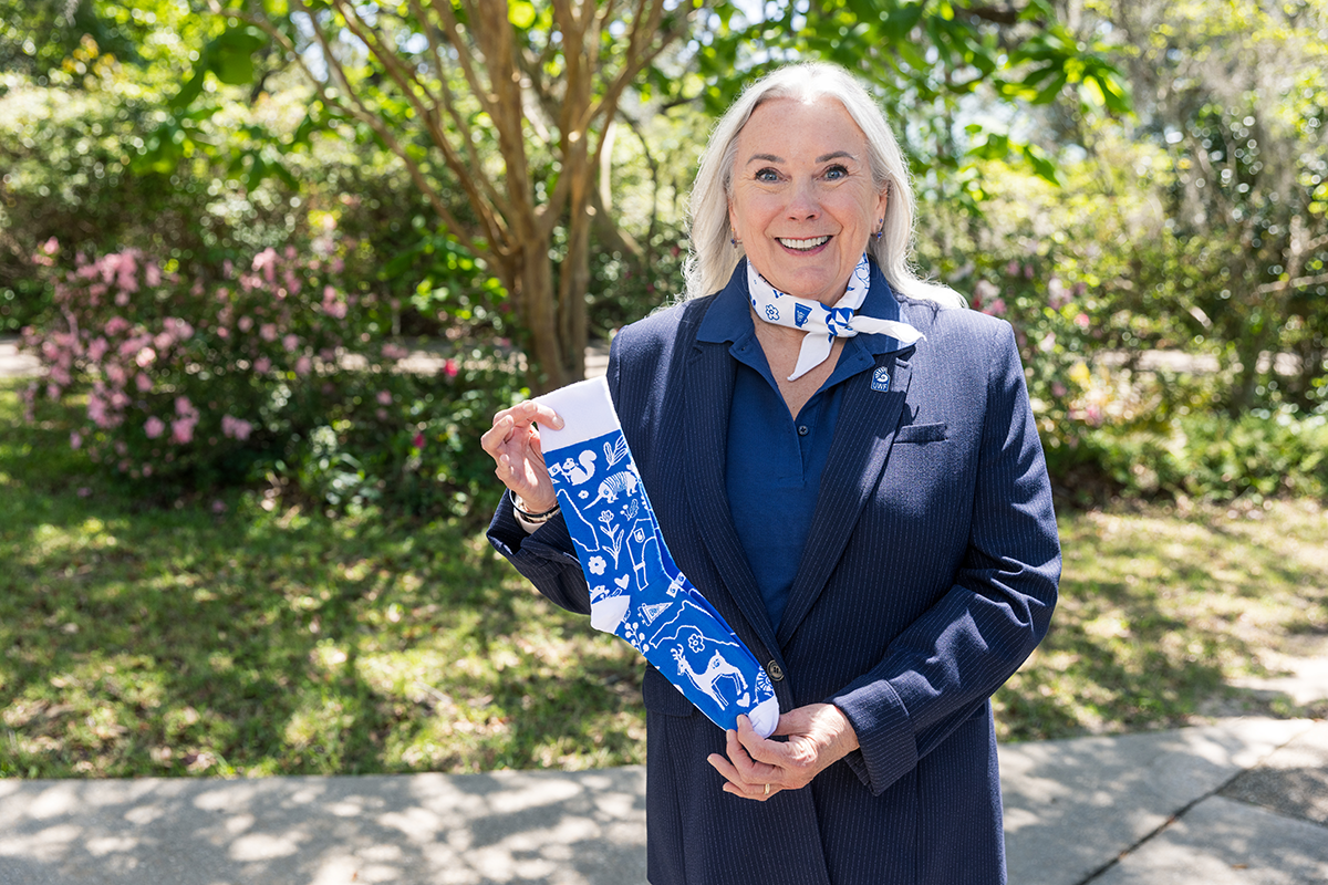 UWF President Martha Saunders stands smiling with a colored-white bandana tied around her neck and holding a colored-blue and white sock in her hands.