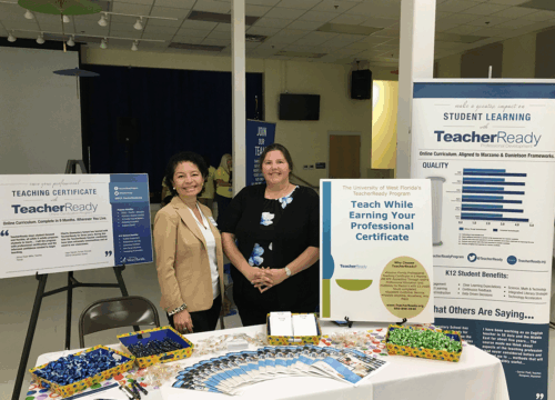 Two people stand behind a table covered with pamphlets about UWF's TeacherReady program in between two tall floor signs