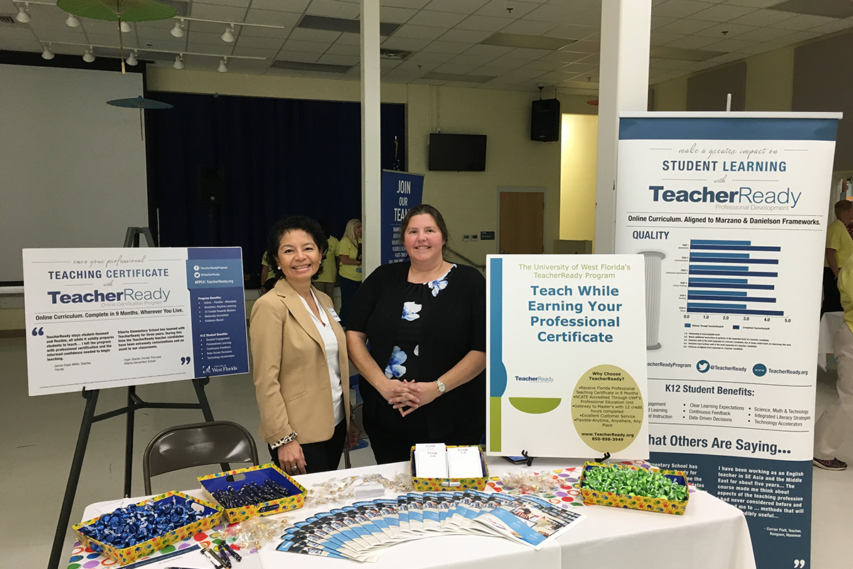 Two people stand behind a table covered with pamphlets about UWF's TeacherReady program in between two tall floor signs