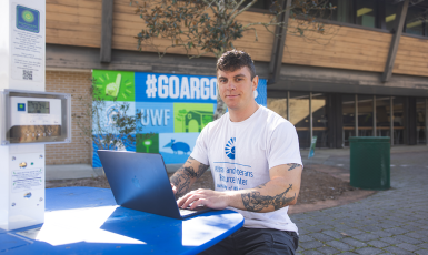 Photo: Man sits at a colored-blue picnic table typing on a laptop in front of a mural colored-blue and green that says #GoArgos