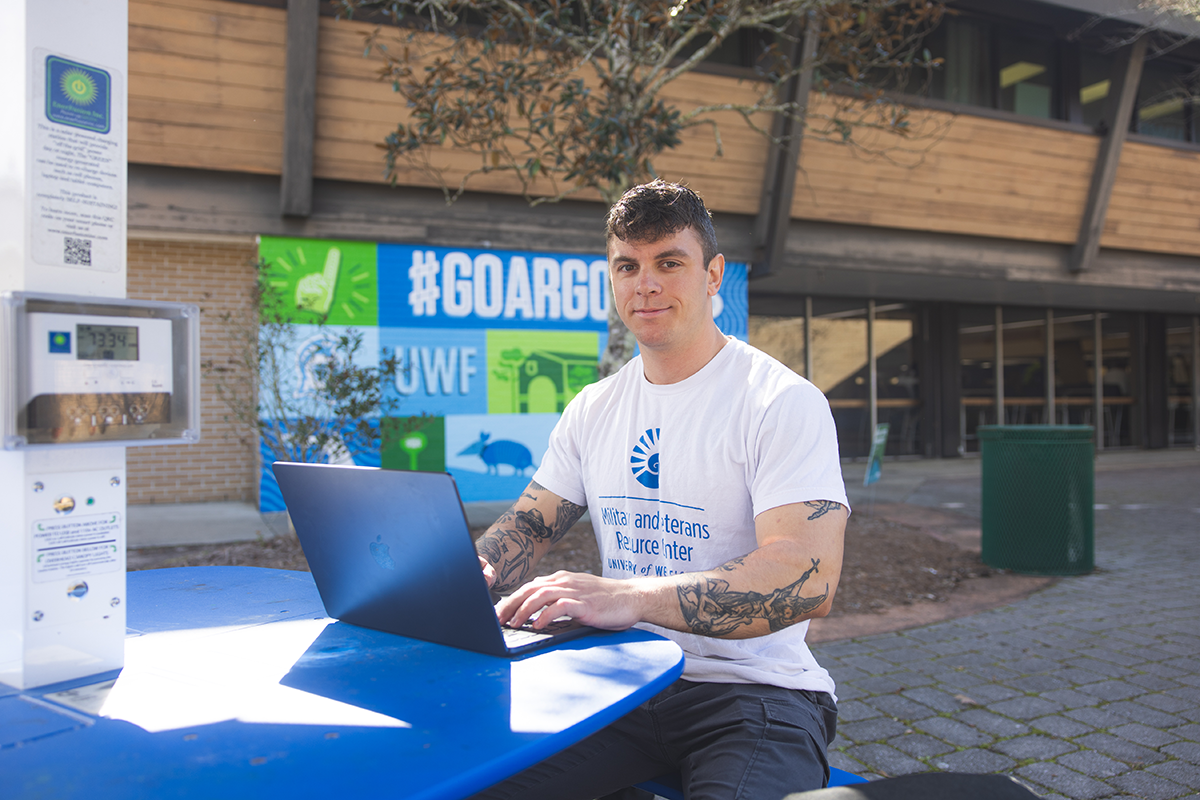 Photo: Man sits at a colored-blue picnic table typing on a laptop in front of a mural colored-blue and green that says #GoArgos