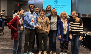 Photo: Eight people stand smiling. One holds a gold trophy. Powerpoint in the background says, "Welcome to the Annual Psi Chi Quiz Bowl"