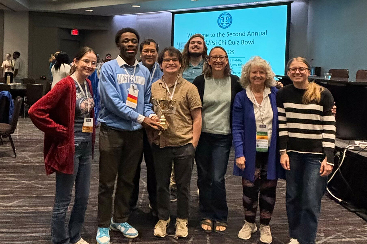 Photo: Eight people stand smiling. One holds a gold trophy. Powerpoint in the background says, "Welcome to the Annual Psi Chi Quiz Bowl"
