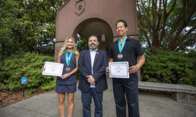 Two students pose next to their professor, holding awards in hand with medals around their necks in front of an archway at the University of West Florida.