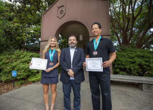Two students pose next to their professor, holding awards in hand with medals around their necks in front of an archway at the University of West Florida.