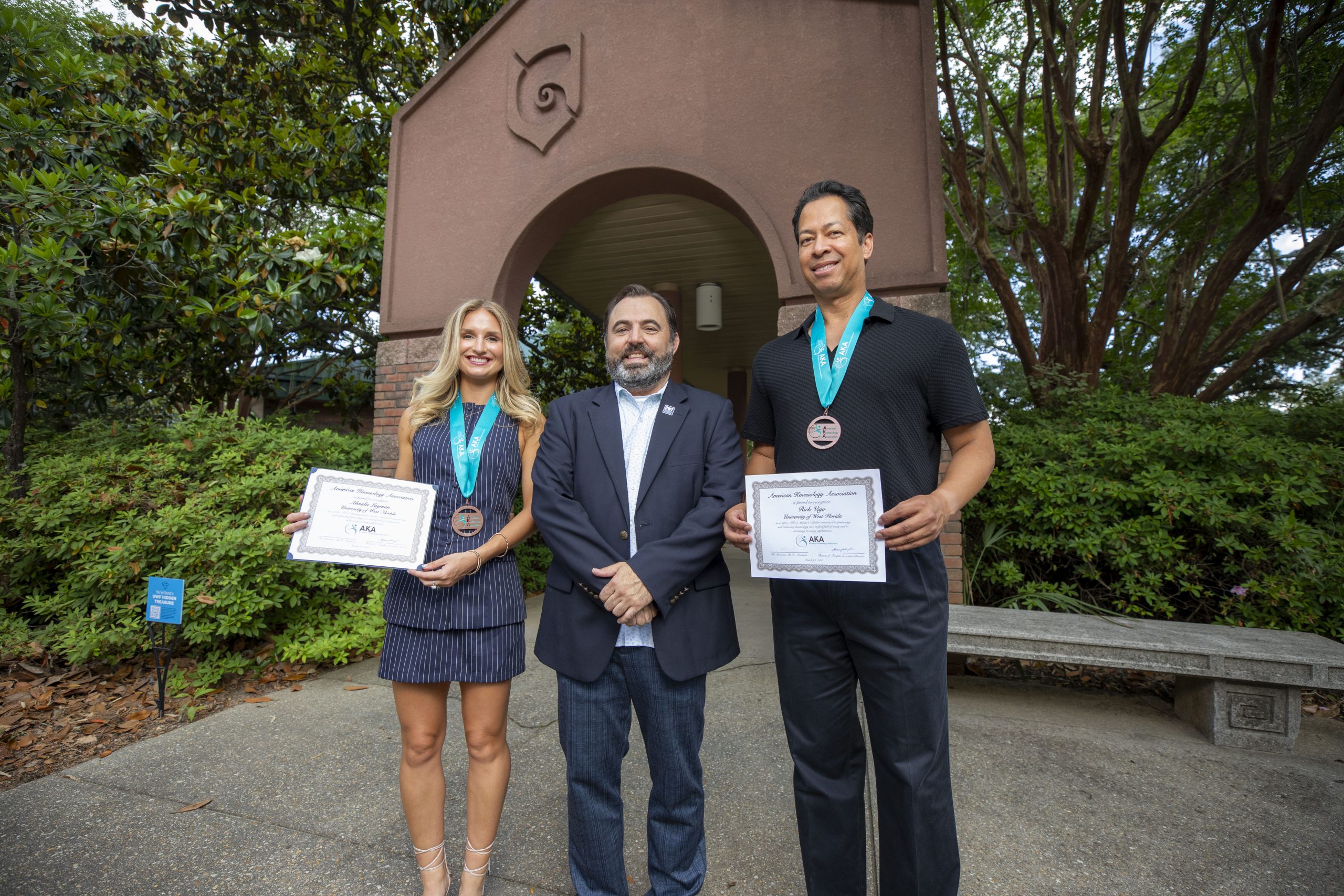 Two students pose next to their professor, holding awards in hand with medals around their necks in front of an archway at the University of West Florida.