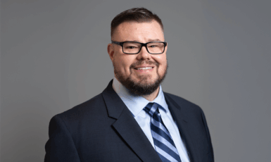 Photo of David Bryant from the shoulders up smiling in a suit and glasses.