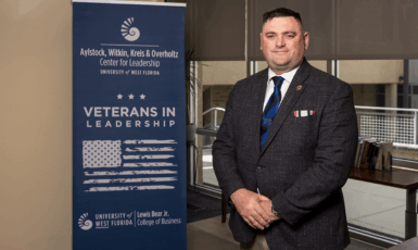 Andrew Del Gaudio stands to the right of a banner that reads, "Aylstock, Witkin, Kreis & Overholtz Center for Leadership. University of West Florida. Veterans in Leadership. University of West Florida. Lewis Bear Jr. College of Business," with an American flag and stars.