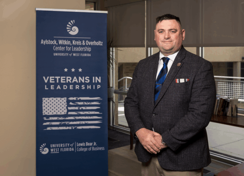 Andrew Del Gaudio stands to the right of a banner that reads, "Aylstock, Witkin, Kreis & Overholtz Center for Leadership. University of West Florida. Veterans in Leadership. University of West Florida. Lewis Bear Jr. College of Business," with an American flag and stars.
