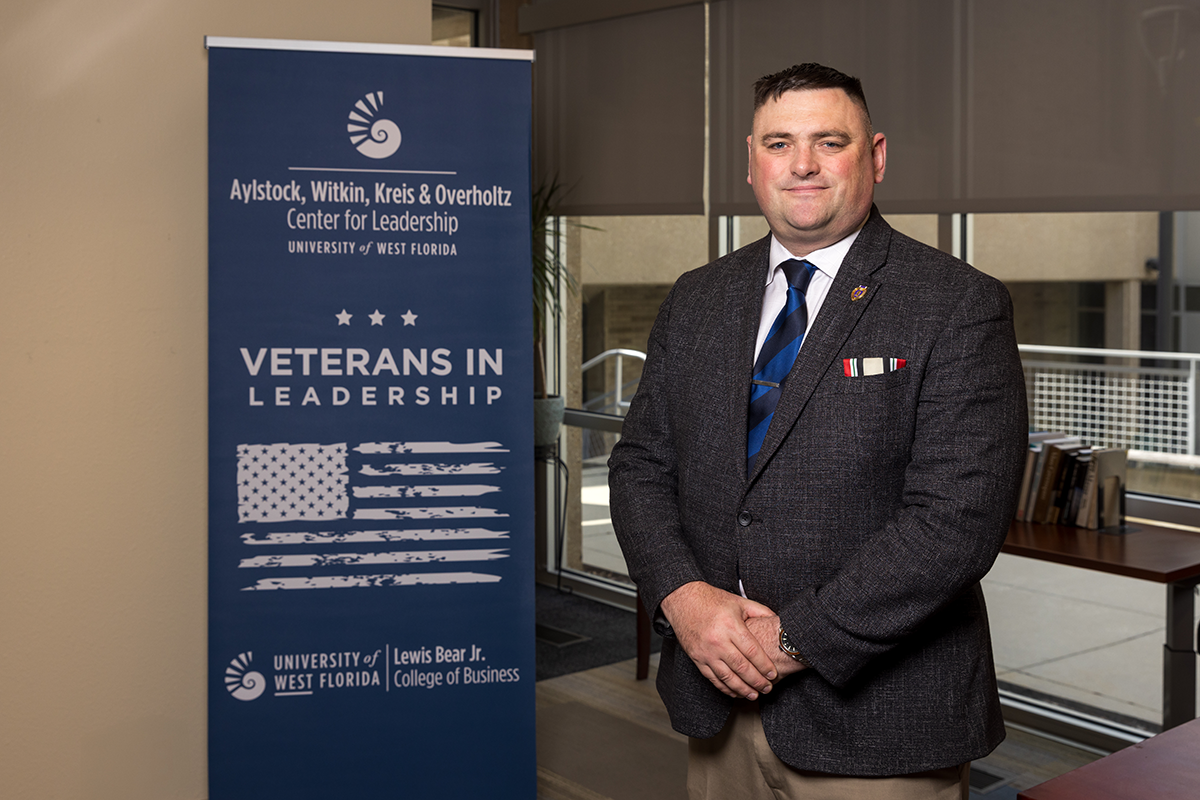 Andrew Del Gaudio stands to the right of a banner that reads, "Aylstock, Witkin, Kreis & Overholtz Center for Leadership. University of West Florida. Veterans in Leadership. University of West Florida. Lewis Bear Jr. College of Business," with an American flag and stars.