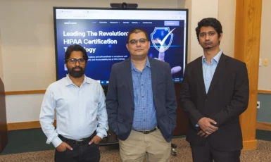 Three people stand in front of a tv screen with a webpage pulled up that reads, "Leading the Revolution: HIPAA Certification Technology"