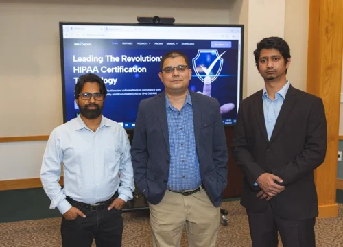 Three people stand in front of a tv screen with a webpage pulled up that reads, "Leading the Revolution: HIPAA Certification Technology"