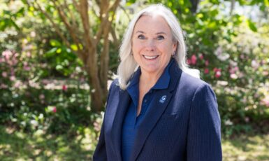 UWF President Martha Saunders smiles with a UWF pin on her jacket in front of greenery