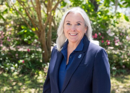 UWF President Martha Saunders smiles with a UWF pin on her jacket in front of greenery