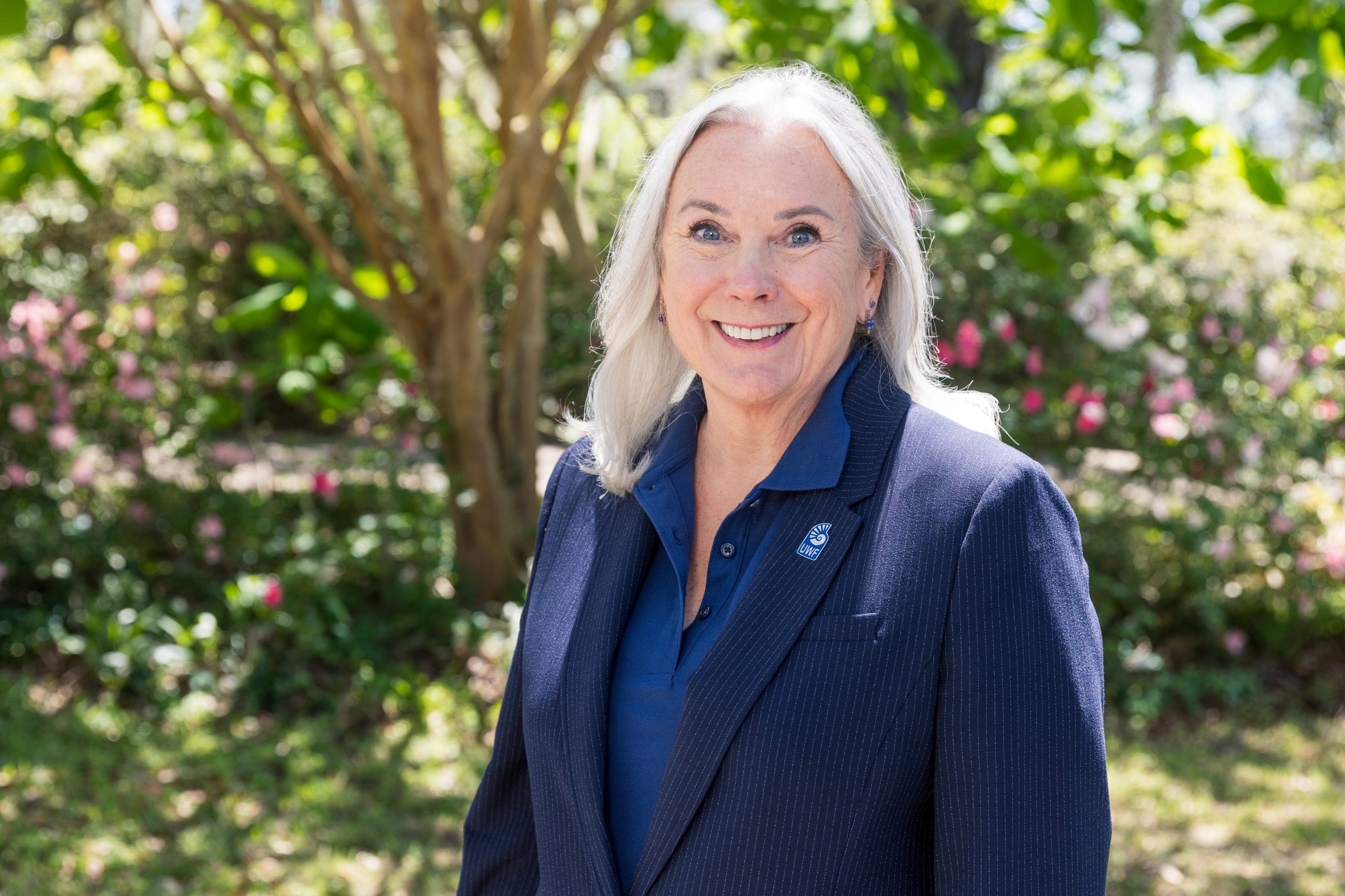 UWF President Martha Saunders smiles with a UWF pin on her jacket in front of greenery