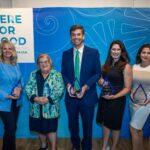 Five award recipients hold their clear glass awards standing in front of a blue backdrop that says, "Here for Good"