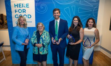 Five award recipients hold their clear glass awards standing in front of a blue backdrop that says, "Here for Good"