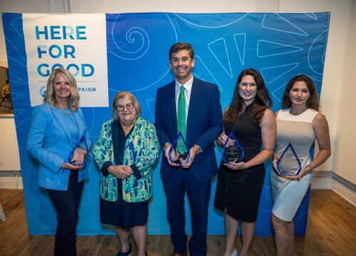 Five award recipients hold their clear glass awards standing in front of a blue backdrop that says, "Here for Good"