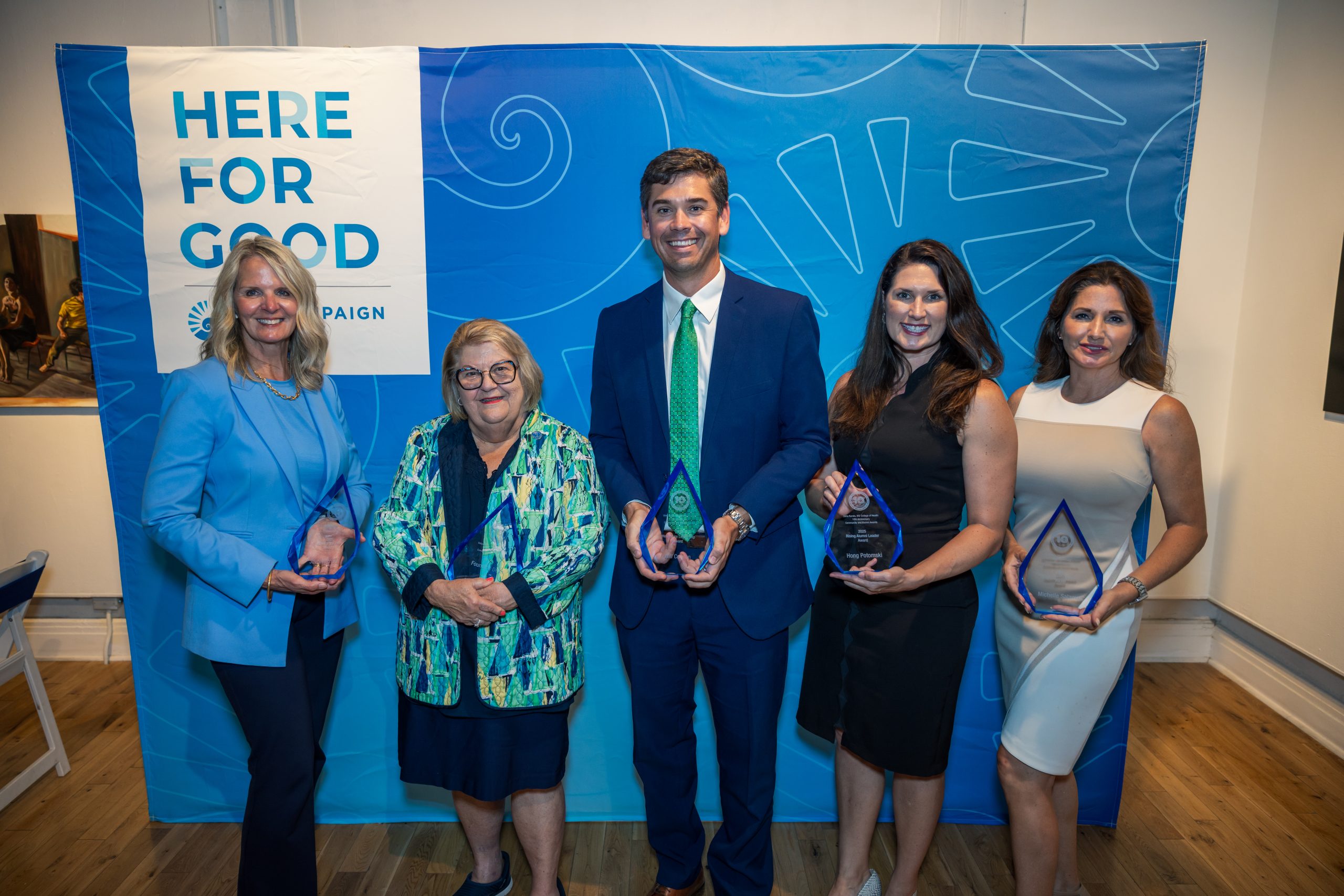 Five award recipients hold their clear glass awards standing in front of a blue backdrop that says, "Here for Good"