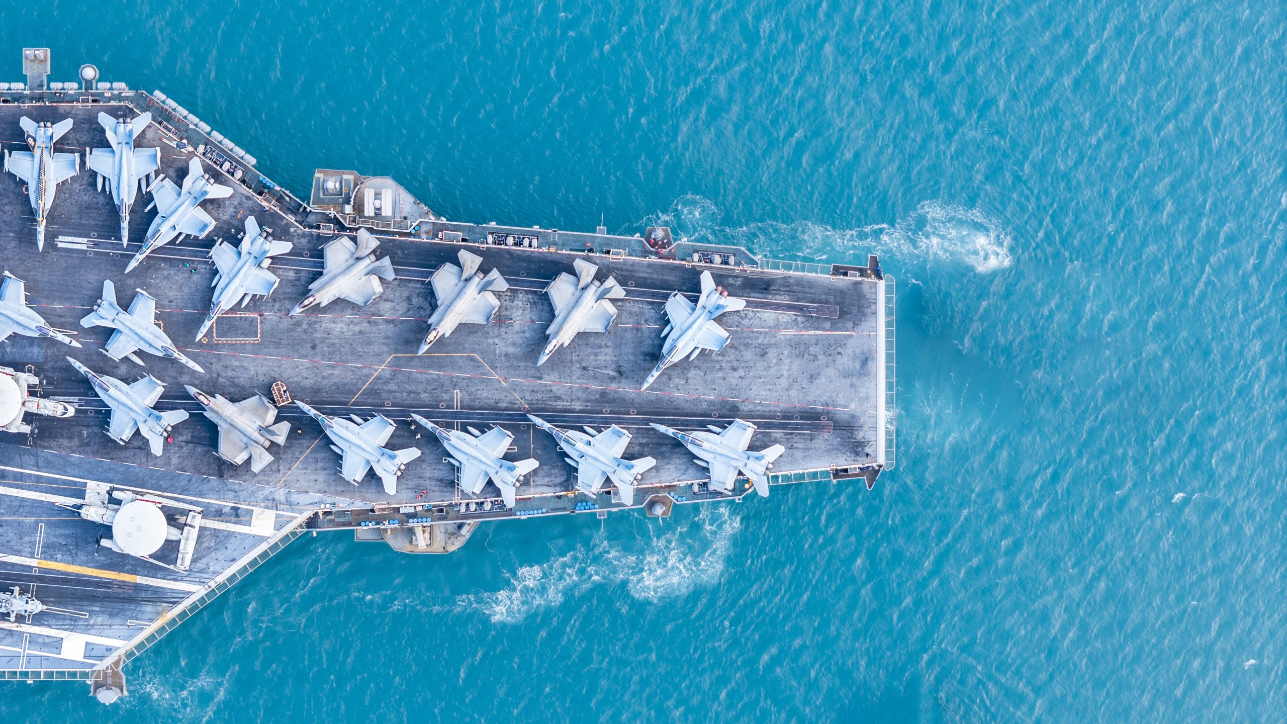Aerial view of a naval aircraft carrier with aircraft on top of it in blue ocean waters.