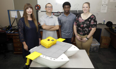A roboboat sits on a table with four students standing behind it.