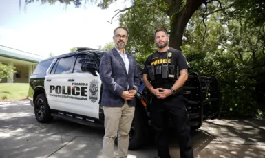 One professor and one police officer stand in front of a Pensacola Police Department cruiser in front of a building and greenery.