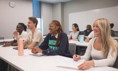 Students laugh in a classroom. Four students sit in the front row, two sit in the middle row, and two are visible in the back row. Text on sweatshirt on student in first row reads, "University of West Florida." Water bottles, notebooks and laptops sit on top of the tables.