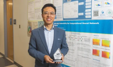 Dr. Shusen Pu stands smiling with his glass award in hand in front of posters of his research.