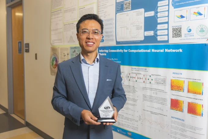 Dr. Shusen Pu stands smiling with his glass award in hand in front of posters of his research.